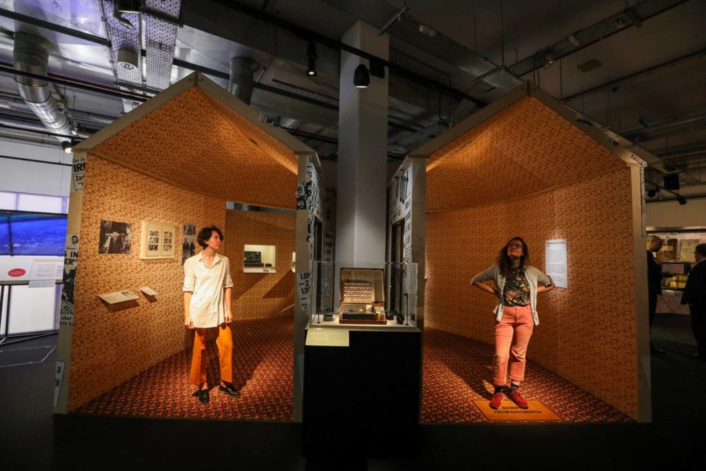 Two people standing in house shaped installations that represent a playful recreation of a 1960s suburban bungalow that was home to Soviet spies.