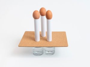 Three eggs balanced in thin cardboard tubes on top of a dining table placement mat on top of three glasses of water
