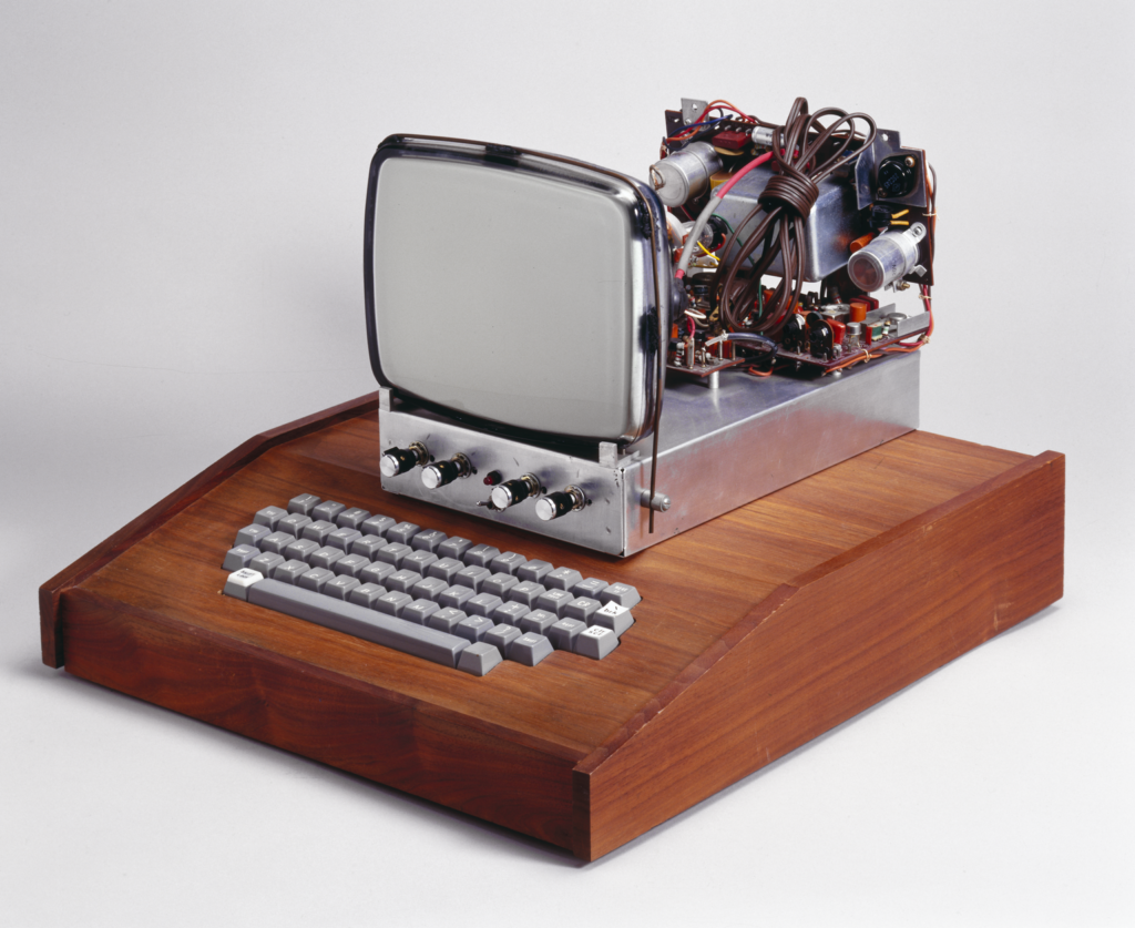 Photograph of the original Apple computer against a light grey background. The screen is small and square, mounted on a metal box with four knobs at the front. The back of the screen is without a case so the motherboard and wires are visible. The screen sits on top of a keyboard made from wood, with light grey plastic buttons. 