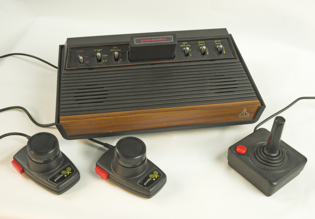 Photograph of the Atari 2600 on a white background. The console itself is made from black plastic, with a wooden panel at the front that has the Atari logo printed on it in white. At the top of the console are multiple switches which read: power (on/off); tv type (color/b.w.); left difficulty (a/b); right difficulty (a/b); game select; game reset. A black plastic cartridge is embedded in the middle of these switches with a label saying “space invaders”. Between the switches and the wooden panel there are grooves in the plastic, and two hollow circles underneath that allow sound to escape from. Connected to the device by black cables are two palm-sized black plastic controllers. Each one has a red plastic button on the side, and a large dial that you can spin. The label underneath says “paddle” and has a logo of two tennis rackets and a ball. On the right-hand side of the photo is another type of controller: this one is square-shaped and slightly larger. There is a small circular red button in the top left corner, and a black plastic stick that can be moved in any direction. 