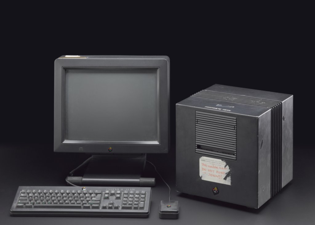 Photograph of the NeXTcube against a black background. The machine consists of a square-shaped screen, a computer keyboard, a computer mouse and a cube-shaped unit. All elements are made from black plastic and feature a small red, yellow and green NeXT logo on them. The cube shapped unit has a scuffed label on the front, with red writing that says: “This machine is a SERVER. DO NOT POWER DOWN!!” 