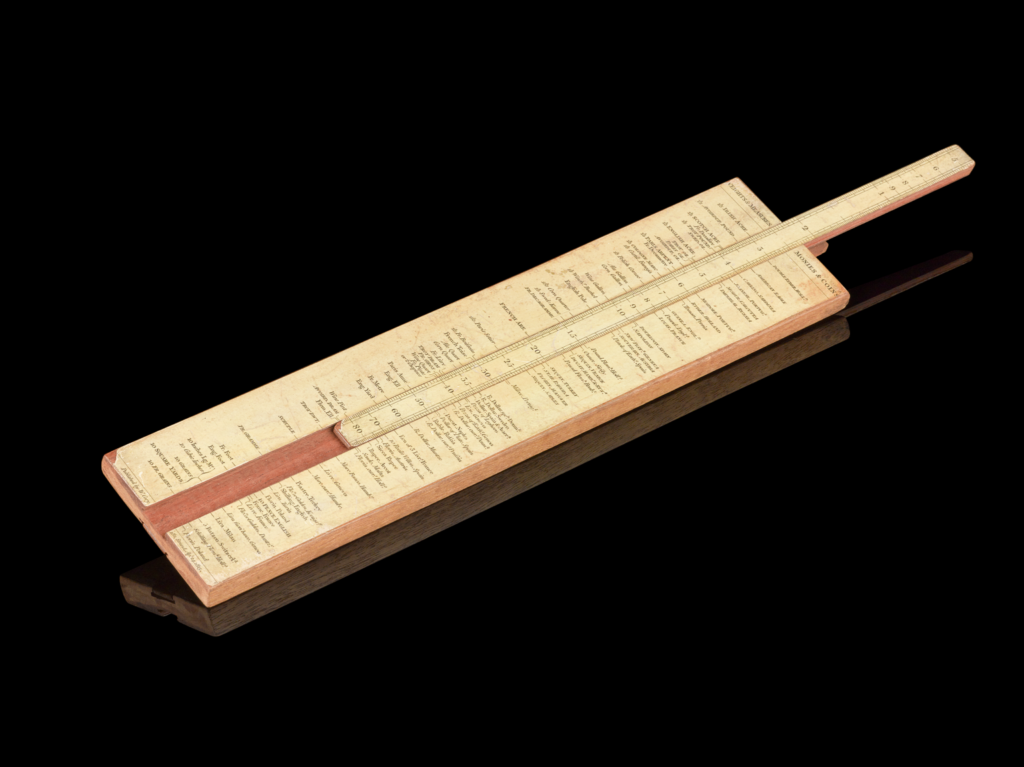Photograph of a slide rule on a black background. It's made out of wood, with yellowed paper on top featuring lots of writing. In the middle there is a strip that can be moved up or down with numbers are etched onto it.