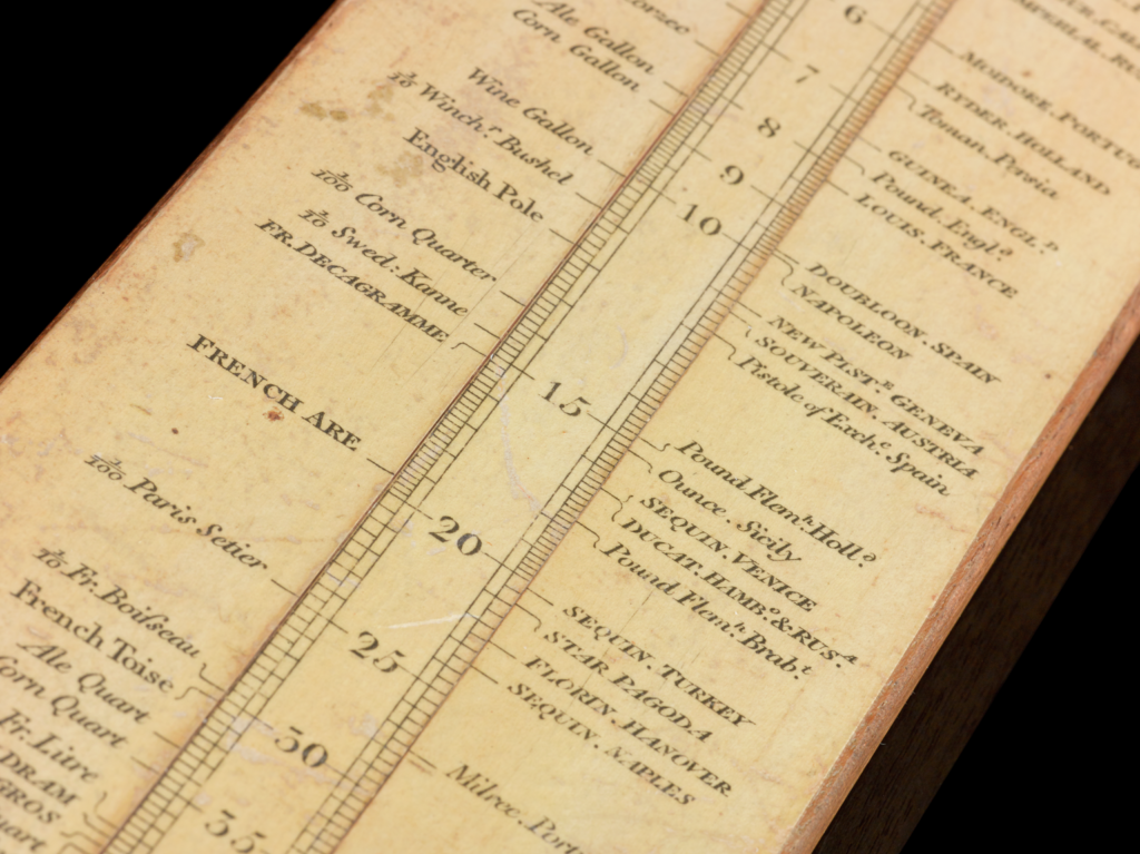 Close up of the slide rule. You can see the middle strip has lines either side of the number to connect it to either side. The number 10 points to "Wine Gallon" on the left, and "NAPOLEON" on the right. 
