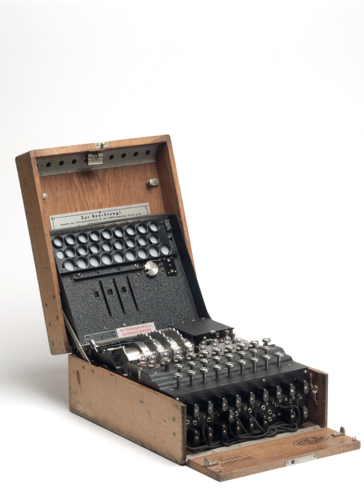 Photograph of an Enigma machine on a white background. It's a square shaped black metal device, encased in light wood. The lid is open, and on the top of the device is a set of three metal wheels, each with the alphabet printed on them. In front of that is a series of small glass bulbs, and a keyboard made up of circular shaped keys. Numbers and letters are printed on the front of the boxc, each with a small socket. Various leads are tangled and plugged into some of these sockets.