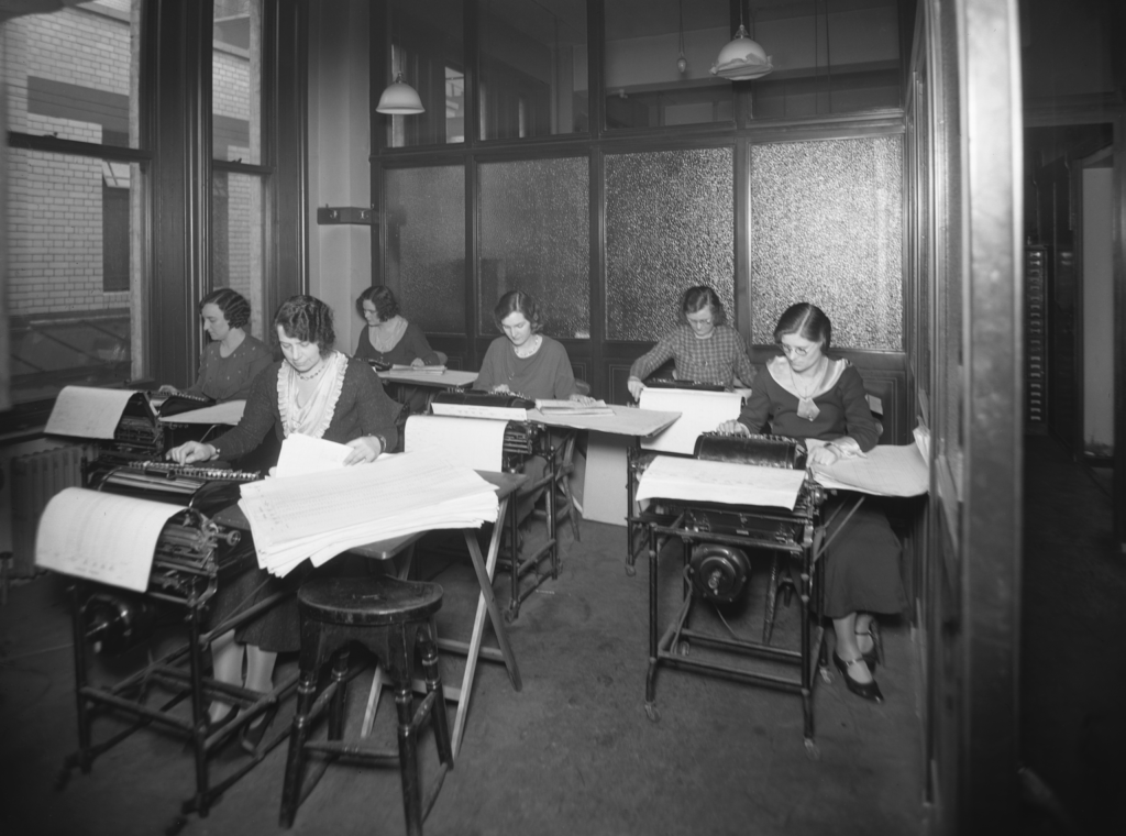 Black and white photograph of six white women working in a small office. Behind them is a frosted glass panel, and to the left is a large window. Each woman sits at an individual machine. These machines have a series of switches and dials on the front, and large sheets of paper at the back. Many of them have more piles of large paper to the side that they are reading. 