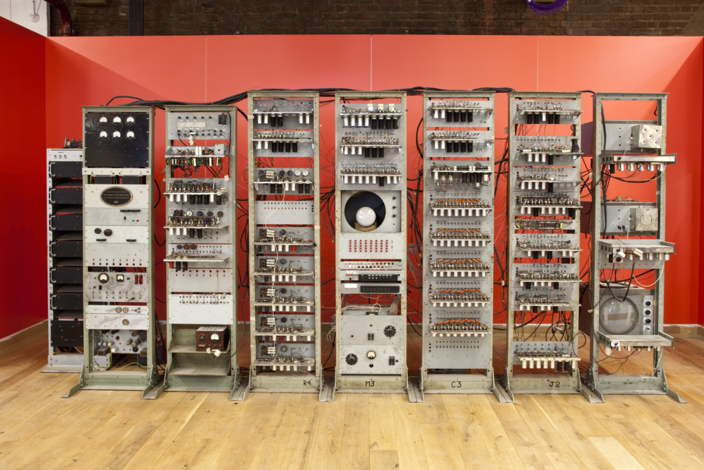 Photograph of the computer ‘Baby’ in a room with a wooden floor and red walls. The computer itself is made up of eight stands, connected by hundreds of black wires, most of which are pulled together at the back. Each stand has multiple sections with various dials, switches, lights and sockets connected by even more wires. 