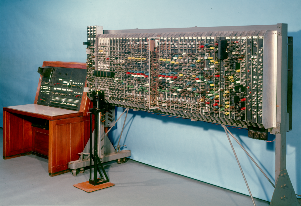 Photograph of the ACE model against a blue background. The machine is in two parts: the first part is a wooden console that is big enough or a person to sit at like a desk. There is a black switchboard embedded into the wood, with white and red buttons. In the top left there is a large viewfinder with small handles either side. Next to it is a large metal frame that holds around 45 vertical strips that are also made of metal. Each strip has various plastic tubes in different colours: black, white, blue, green, red and yellow. One of the strips is pulled out to show that the tubes are connected by an an intricate maze of wiring. 