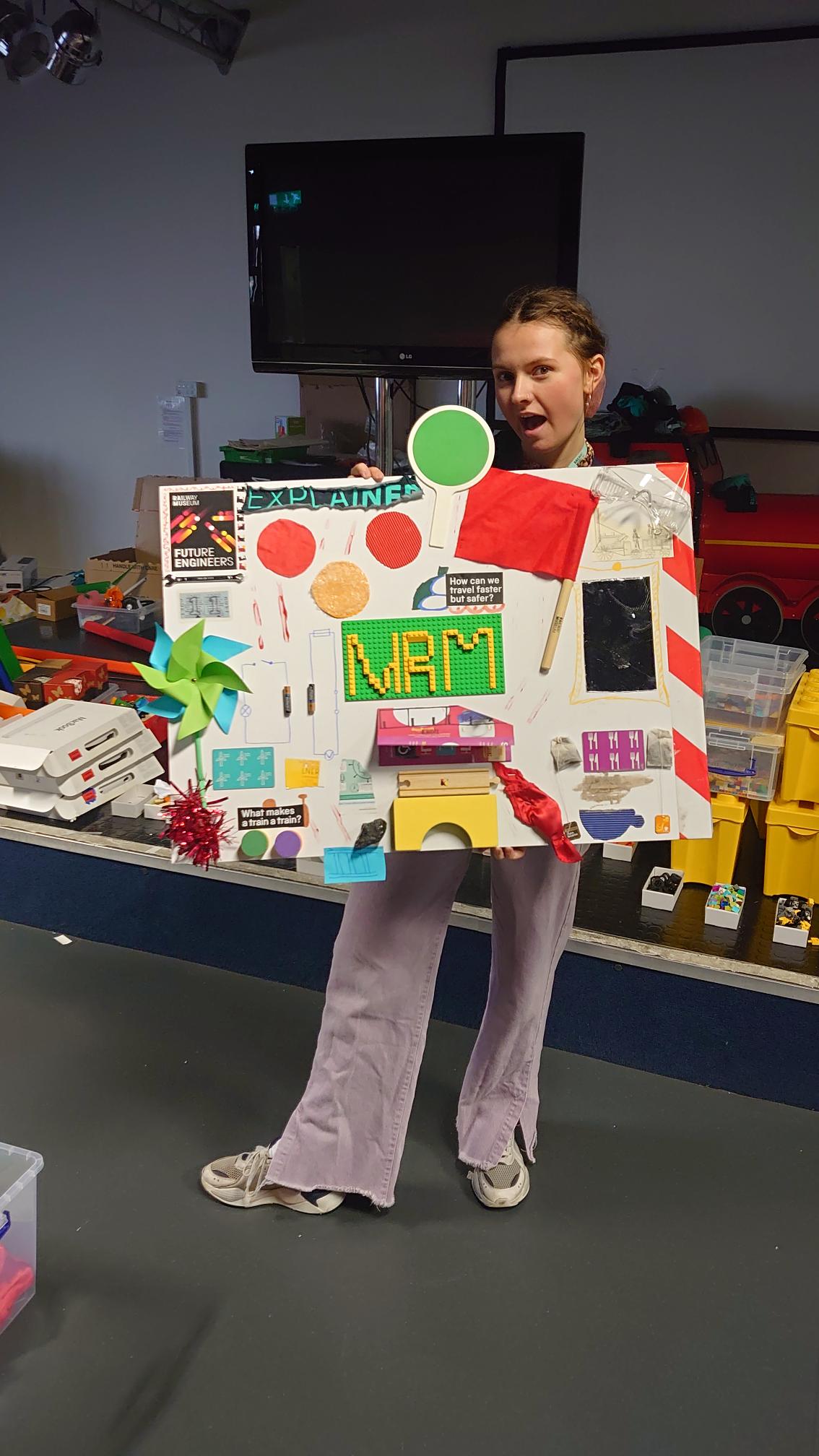 A young woman wearing purple trousers and her hair in plaits. She is holding an NRM Sensescape, a A1 moodboard with lego, bridges, paper windmills, tea bags, and flags