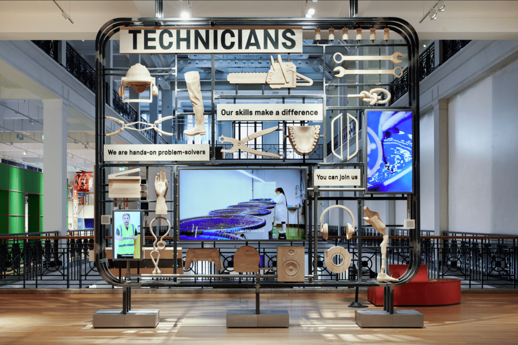 View entering the technicians gallery at the science museum. A floor to ceiling steal display holds large replicas of tools used by technicians. The display is designed to resemble an air fix model kit. Two screens display film clips of technicians at work while one displays a video of a British sign language interpreter.