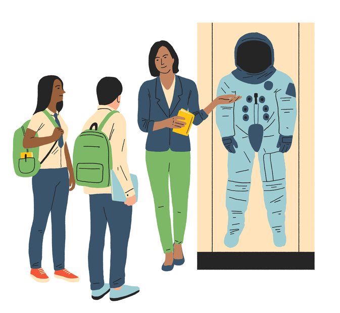Two school students dressed in blue trousers and white tops listen to a teacher explaining about an astronaut space suit.
