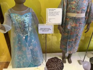 A blue and sparkly children dress with a snowflake cape, positioned next to a small rectangular plaque with the question “What would be your dream outfit to express who you are?” written on it.