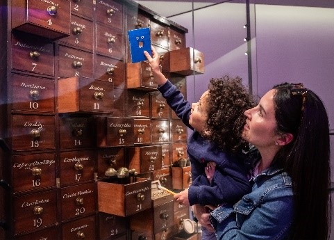 A young visitor, held in a grown ups arms, holds a blue square shape with google eyes up to a glass cabinet. Inside the cabinet is a large wooden "draw like" object with multiple square shaped drawers. 